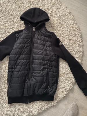 Svart pufferjacka med huva från Moncler - Säljer en svart pufferjacka från Moncler med huva och quiltad front. Jackan har släta ärmar, två dragkedjefickor framtill och ribbade muddar. Moncler-logga på ärmen ger en exklusiv touch. Perfekt för kalla dagar och riktigt snygg till streetwear.