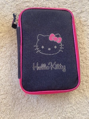 Hello Kitty svart pennfodral - Snyggt marinblått pennfodral från Hello Kitty med rosa detaljer och dragkedja. Framsidan har ett glittrigt Hello Kitty-tryck och en rosa rosett. Perfekt för att hålla ordning på pennor och småprylar i väskan.