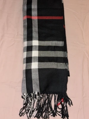 Svart halsduk med rutigt mönster - Snygg svart halsduk med klassiskt rutigt mönster i vitt, grått och en röd linje. Halsduken har fransar i ändarna och är tillverkad i ett mjukt tyg som känns skönt mot huden. Perfekt accessoar för att lyfta din outfit.