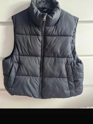 Svart dunväst med hög krage - Snygg svart dunväst med hög krage och dragkedja framtill. Västen har quiltad design och två sidofickor. Perfekt att slänga över en hoodie eller tröja för extra värme. Materialet är mjukt och puffigt, vilket ger en cool och avslappnad look.
