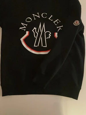 Svart Moncler tröja med stor logga - Svart hoodie från Moncler med stor vit och röd logga på bröstet samt Moncler-märke på ärmen. Klassisk passform och mjukt material, perfekt för en stilren och trendig look.