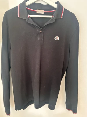 Svart långärmad polotopp från Moncler - Klassisk svart långärmad polotopp från Moncler med vit och röd rand på kragen. Toppens material är bomull och den har en liten Moncler-logga på bröstet. Perfekt för en stilren och sportig look.