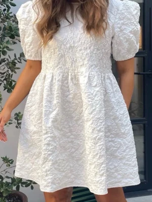 Vit kort klänning med puffärm - Supersöt vit kortklänning med puffärmar och strukturmönstrad yta. Klänningen har rund halsringning och en lätt utställd kjol. Perfekt för dig som gillar en feminin och trendig look. Tillverkad i ett mjukt tyg som känns bekvämt mot huden. Klänningen är insydd i midjan och axlarna är nerkortade en del. Därav är storleken närmare ”S” även om den är köpt i ”M”. Använd bara på studenten så inprincip helt ny.
