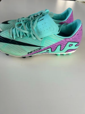 Nike Mercurial Air Zoom fotbollsskor - Snygga Nike Mercurial Air Zoom fotbollsskor i mintgrönt och lila med svart swoosh. Skorna har snörning, låg profil och greppvänlig dobbsula för bästa fäste på planen. Tillverkade i syntetmaterial som ger lätt känsla och bra bollkontroll. Perfekta för dig som vill sticka ut på planen.