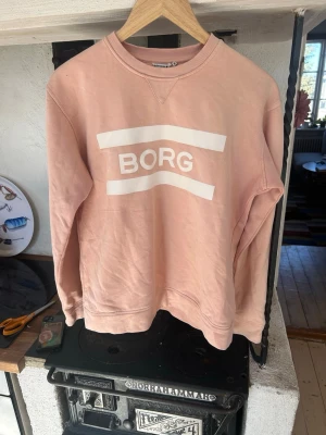 Ljusrosa sweatshirt från BORG - Ljusrosa sweatshirt från BORG med vit logga framtill. Tröjan har rund halsringning, ribbade muddar vid ärmslut och nederkant samt en avslappnad passform. Perfekt för en casual och trendig look.