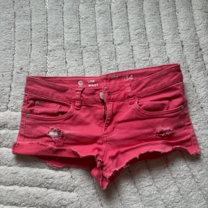 Rosa low waist shorts med slitningar - Säljer ett par rosa low waist shorts från Cubus i storlek 34. Shortsen har slitna detaljer och råa kanter för en avslappnad look. Klassisk femficksmodell med knapp och dragkedja framtill. Perfekta för varma dagar!