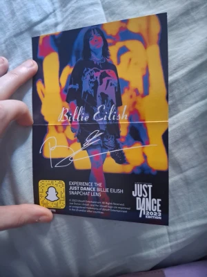 Just Dance 2023 Billie Eilish kort - Unikt samlarkort från Just Dance 2023 Edition med Billie Eilish-tema. Ena sidan har en färgstark bild av Billie Eilish och info om Snapchat-lins, andra sidan ser ut som ett kassettband med låten 'Therefore I Am'. Perfekt för fans av spelet och artisten.