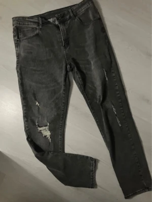 Grå slitna skinny jeans Manfinity  - Säljer ett par grå skinny jeans med slitningar och hål på benen för en trendig look. Jeansen har klassisk femficksmodell och hög midja. Perfekta för dig som gillar en avslappnad och edgy stil.