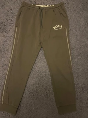Gröna boss byxor  - Snygga olivgröna mjukisbyxor från BOSS med dragsko i midjan och ribbade muddar vid bensluten. Byxorna har sidofickor, en bakficka och diskreta beigea ränder längs benen. Tillverkade i mjukt material för maximal komfort. Perfekta för en avslappnad stil. Storlek s skriv om ni är intresserade 