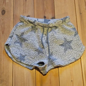 Grå fluffiga shorts med stjärnor - Supermysiga loungewear-shorts i grått teddy-material med stora stjärnor i en mörkare grå nyans. Shortsen har resårmidja med dragsko för skön passform och rundade benslut. Perfekta för chill hemma eller mysiga kvällar.