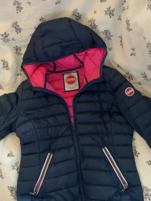 Mörkblå Colmar pufferjacka med huva - Snygg mörkblå pufferjacka från Colmar med huva och rosa foder. Jackan har två dragkedjeförsedda fickor framtill och Colmar-logga på ärmen. Perfekt för lite varmare vår dagar🌸 riktigt skön att bära. Kan fixa upp en outfit så lätt!