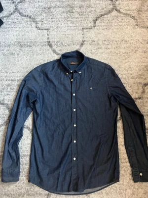 Mörkblå skjorta från Morris - Snygg mörkblå skjorta från Morris med button-down krage och vita knappar. Skjortan har lång ärm och en liten broderad logga på bröstet. Tillverkad i ett mjukt bomullstyg som känns bekvämt och stilrent.