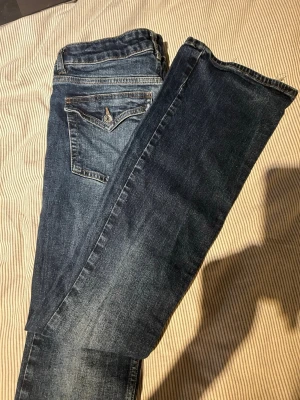 Blå bootcut jeans med fickdetalj - Säljer ett par klassiska blå bootcut jeans med snygg fickdetalj bak och knappstängning. Jeansen har en lätt tvättad look och raka ben som går ut lite nedtill. Perfekta till sneakers eller boots för en avslappnad stil.