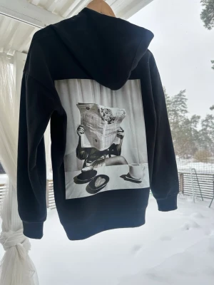 Svart hoodie med ett unikt retro trygg på ryggen  - Svart hoodie med stor svartvit print på ryggen som visar en person som läser tidningen vid ett frukostbord. Klassisk känguruficka framtill och ribbade muddar. Perfekt för dig som gillar streetwear och unika detaljer. Materialet känns mjukt och bekvämt.