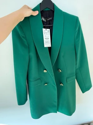 Lång Grön kavaj från Zara med guldknappar - Stilren grön lång kavaj/klänning  från Zara med dubbelknäppning och snygga guldknappar framtill. Klassisk krage och två fickor. Perfekt för dig som vill sticka ut med färg och samtidigt hålla det elegant. Materialet är polyester.