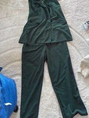 Mörkgrön ärmlös jumpsuit med knappar - Stilren mörkgrön jumpsuit med vida ben och ärmlös design. Framtill finns dubbla rader med knappar och två fickor. Materialet är mjukt och faller snyggt. Perfekt för dig som vill ha en cool och avslappnad look.
