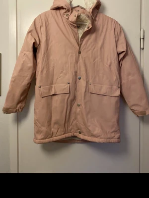 Ljusrosa regnjacka med huva - Snygg ljusrosa regnjacka med mysigt foder i huvan och stora fickor framtill. Jackan har tryckknappar, dragsko i huvan och  reflexdetalj baktill. Perfekt för kyliga dagar och ger en avslappnad streetstyle-look.