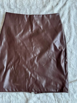 Brun skinnimitation kjol med slits - Snygg brun kjol i skinnimitation med rak passform och slits bak. Kjolen har en stilren look och stängs med dragkedja bak. Perfekt att matcha med en enkel topp eller stickad tröja.