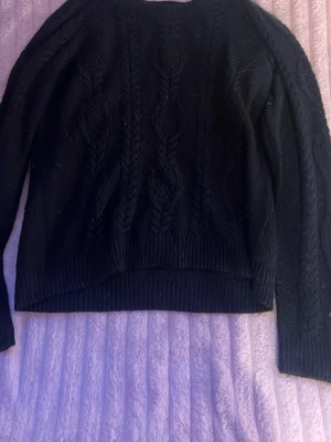 Svart kabelstickad tröja - Mysig svart kabelstickad tröja med rund hals och långa ärmar. Tröjan har ett klassiskt flätmönster framtill och ribbade muddar vid ärmslut och nederkant. Perfekt för kyligare dagar och enkel att matcha med olika outfits.