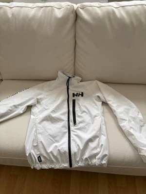Vit vindjacka från Helly Hansen - Snygg vit vindjacka från Helly Hansen med svart dragkedja och logga på bröstet. Jackan har hög krage, elastisk nederkant och en bröstficka med dragkedja. Perfekt för blåsiga dagar och enkel att matcha med olika outfits.