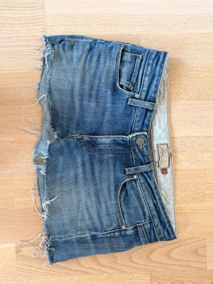 Blå jeanskjol från Paige, kort modell - Säljer en kort jeanskjol från Paige i klassisk blå denim. Kjolen har råa, fransiga kanter och slitna detaljer för en avslappnad look. Femficksmodell med knapp och dragkedja framtill. Perfekt för dig som gillar en edgy och trendig stil.