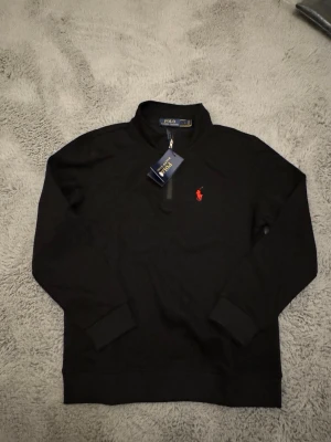 Svart halvzip tröja från Polo Ralph Lauren - Svart långärmad tröja från Polo Ralph Lauren med halv dragkedja och röd broderad logga på bröstet. Tröjan har ribbade muddar vid ärmslut och nederkant, samt en klassisk och stilren design som passar till mycket. (Tröjan är L i barnstorlek vilket är ungefär S/xs i herr storlek)
