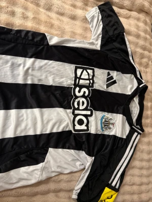 Newcastle United matchtröja Adidas - Säljer en svartvit randig Newcastle United matchtröja från Adidas med klubbmärke och sponsorlogga på bröstet. Tröjan har korta ärmar, klassiska Adidas-ränder på axlarna och gul kaptensbindel på ena ärmen. Perfekt för fotbollsfans!