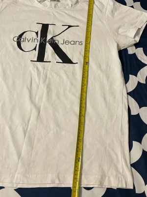 Vit Calvin Klein Jeans t-shirt - Snygg vit t-shirt från Calvin Klein Jeans med stort svart CK-logotyptryck på framsidan. Klassisk rund hals och korta ärmar. Tillverkad i mjukt bomullsmaterial som känns skönt mot huden. Perfekt för en clean och stilren look.