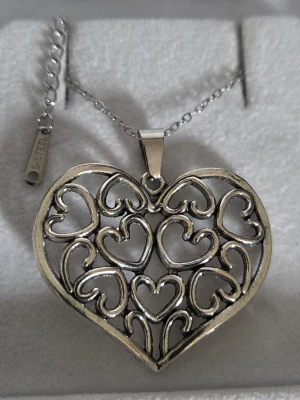 Halsband med hjärthänge i stål - Snyggt halsband i silverfärgat rostfritt stål med ett stort hjärtformat hänge. Hänget har ett öppet, detaljerat mönster med flera mindre hjärtan inuti. Kedjan är tunn och justerbar med en bricka märkt 'S. Steel'. Perfekt för dig som gillar romantiska detaljer. Kedjan är 45 cm och hänget ca 3 cm.