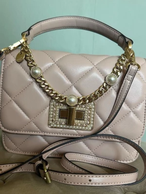 Beige quiltad handväska från ALDO - Snygg beige quiltad handväska från ALDO med guldfärgade detaljer och pärlprydd kedja. Väskan har ett dekorativt spänne framtill med glittrande stenar och ett justerbart axelband. Tillverkad i konstläder med både handtag och axelrem för olika stilar.