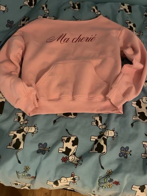 Rosa hoodie med Ma chérie-tryck - Ljusrosa hoodie med texten 'Ma chérie' i snygg skrivstil på bröstet. Hoodien har en klassisk känguruficka framtill och ribbade muddar vid ärmslut och nederkant. Tillverkad i mjukt sweatshirtmaterial, perfekt för en chill och trendig look.