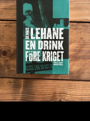 En drink före kriget - Dennis Lehane - En spännande thrillerbok med hård pärm i svart och grönt, skriven av Dennis Lehane. Boken har tydlig, stor vit text på omslaget och handlar om privatdetektiverna Patrick Kenzie och Angie Gennaro i Boston.