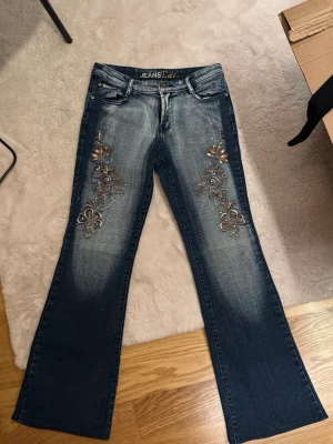Blå bootcut jeans med blommig brodyr - Snygga blå bootcut jeans från JEANS Edi med detaljerad blommig brodyr i brons och beige på båda benen. Jeansen har klassisk femficksmodell, slitningar framtill och normalhög midja. 