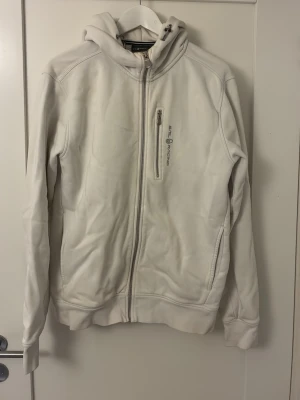 Vit hoodie från Sail Racing - Säljer en vit Sail Racing bowman med dragkedja framtill och en bröstficka med logga. Mjuk fleece på insidan och huva med dragsko. Perfekt för chill dagar eller när du vill ha en clean look.