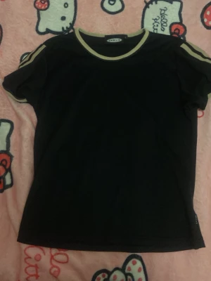 Svart t-shirt med beige detaljer V-RUS - Svart t-shirt från V-RUS med beige ribbade detaljer vid halsringning och ärmslut. Klassisk passform och korta ärmar, perfekt för en avslappnad look. T-shirten har sportiga ränder på axlarna och är tillverkad i mjukt material.