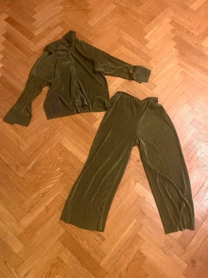 Olivgrönt set i glansigt tyg - Säljer ett olivgrönt loungewear-set med skjorta och vida byxor i glansigt, ribbat material. Skjortan har långa ärmar, breda manschetter och krage. Byxorna har resår i midjan och avslappnad passform. Perfekt för chill dagar hemma.Skriv gärna för mått! 💕