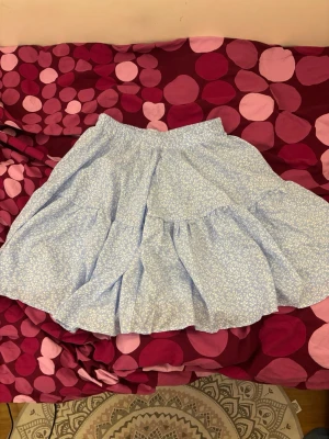 Ljusblå blommig volangkjol - Supersöt ljusblå kjol med vitt blommönster och volangdetaljer. Kjolen har en luftig och flowy passform, perfekt för varma dagar. Resår i midjan som är stretchig och bekväm. Passar perfekt till en enkel topp eller stickad tröja.