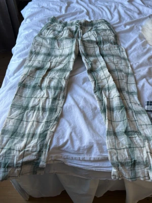Rutiga pyjamasbyxor i grönt och vitt - Säljer ett par lösa och bekväma pyjamasbyxor med grönt och vitt rutmönster. Byxorna har resår i midjan och är gjorda i ett mjukt, tunt tyg som känns skönt mot huden. Perfekta för chillkvällar hemma eller när du bara vill ta det lugnt.