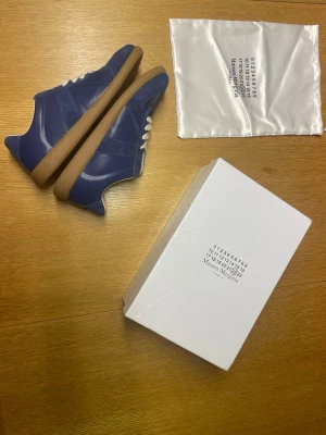 Blå Maison Margiela sneakers - Snygga blå sneakers från Maison Margiela med klassisk gummisula och vita skosnören. Skorna är tillverkade i en mix av läder och mocka, vilket ger en schysst kontrast i material och finish. Perfekta för dig som vill ha något unikt och stilrent i garderoben.