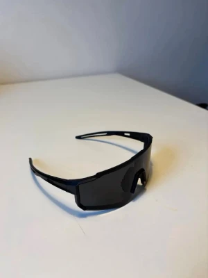 Pitwipers - Säljer ett par svarta solglasögon med bred, rektangulär sköldformad lins och sportig design. Bågarna är i svart plast och glasen är mörkt tonade, perfekta för en cool och modern look. Passar dig som gillar futuristisk stil.