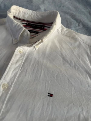 Vit kashmere skjorta från Tommy Hilfiger - Klassisk vit skjorta från Tommy Hilfiger med knappkrage och broderad logga på bröstet. Tillverkad i en mjuk blandning av bomull och kashmir för extra komfort. Skjortan har långa ärmar och stilrena knappar framtill.