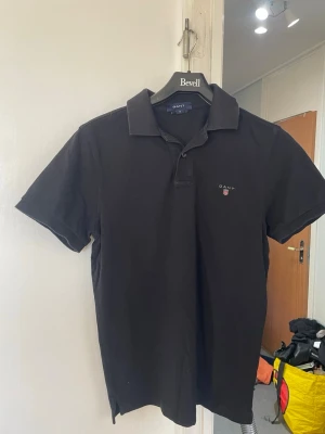 Svart pikétröja från Gant - Svart pikétröja från Gant i regular fit med kort ärm och klassisk krage. Tröjan har en liten broderad logotyp på bröstet.den mörkare i verkligheten,på bild ser liten gråaktig,så den är mörkare  och den är svart .på bild ser liten gråaktig,så den är mörkare  och den är svart