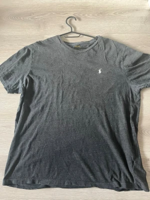 Mörkgrå t-shirt från Polo Ralph Lauren - Snygg mörkgrå t-shirt från Polo Ralph Lauren med klassisk rund hals och korta ärmar. T-shirten har den ikoniska broderade loggan i vitt på bröstet och är tillverkad i mjuk bomull för en skön känsla.