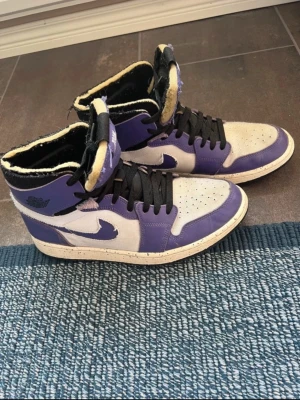  Nike Air Jordan 1 High - Säljer ett par Nike Air Jordan 1 High sneakers i lila och vitt med svarta detaljer. Skorna har klassisk hög siluett, svart snörning och synlig Nike-logga på sidan. Ovandelen är i läder och sulan är vit med svart yttersula. Perfekta för dig som gillar streetwear och vill sticka ut.