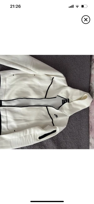 Vit Nike vindjacka med svarta detaljer - Nike vindjacka i vitt med svarta kontrastdetaljer och dragkedja. Jackan har huva, lång ärm och sportig look. Materialet är polyester och jackan har en clean design med Nike-logga på bröstet. Perfekt för dig som gillar streetwear och sportig stil.