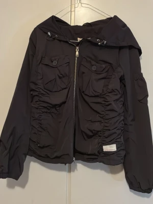 Svart Molly vindjacka med huva och fickor - Svart of Molly vindjacka med huva, två bröstfickor med knappar och dragkedja framtill. Jackan har rynkade detaljer på sidorna och en liten patch nertill. Tillverkad i syntetmaterial och har långa ärmar.