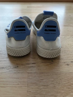 Adidas Pharrell Williams vita sneakers - Snygga vita sneakers från Adidas Pharrell Williams med blå detaljer på häl och plös. Säljs utan snören. Innersulor i original medföljer.