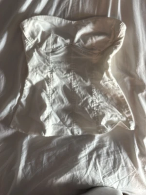 Vit axelbandslös korsettopp - Säljer en stilren vit korsettopp med axelbandslös design och markerad byst. Toppen har en figurnära passform och lätt glansigt tyg som ger en elegant look. 