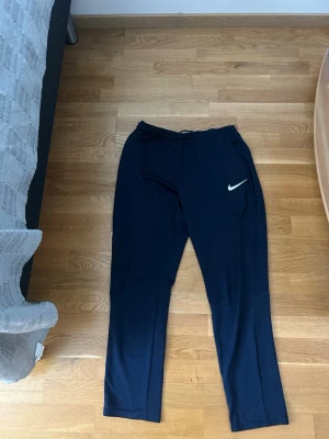 Mörkblåa träningsbyxor från Nike - Säljer ett par mörkblåa träningsbyxor från Nike med vit logga på vänster ben. Byxorna har resår i midjan och raka ben. Perfekta för gymmet eller löpning. Tillverkade i ett lätt och stretchigt material som andas.