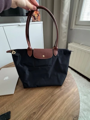 Svart Longchamp Le Pliage handväska - Stilren svart handväska från Longchamp Le Pliage med bruna läderhandtag och lock. Väskan är tillverkad i nylon med detaljer i brunt läder och har guldiga metalldetaljer. Perfekt storlek för att bära med sig allt man behöver.